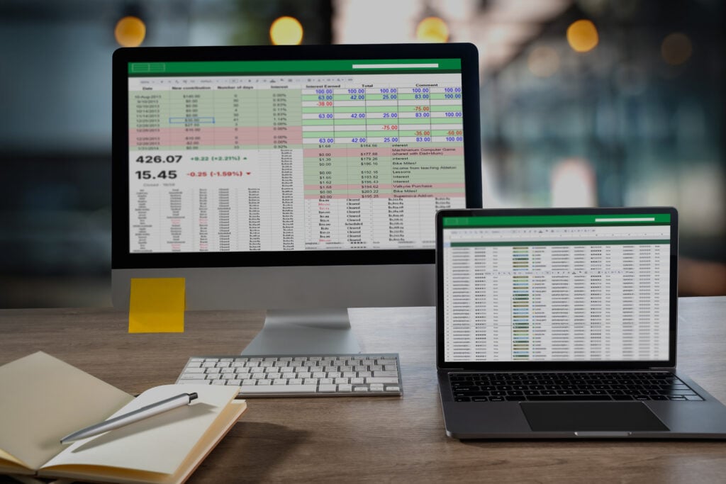 Desktop monitor and laptop display a detailed spreadsheet with financial figures and color coded rows on a tidy office desk ideal visual for analytics KPI