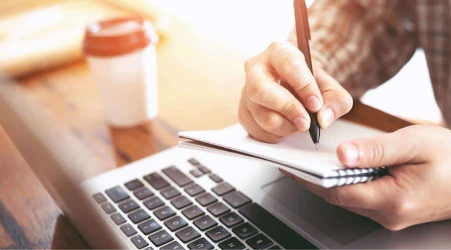 A person writing in a notebook with a laptop and coffee cup on a wooden table.