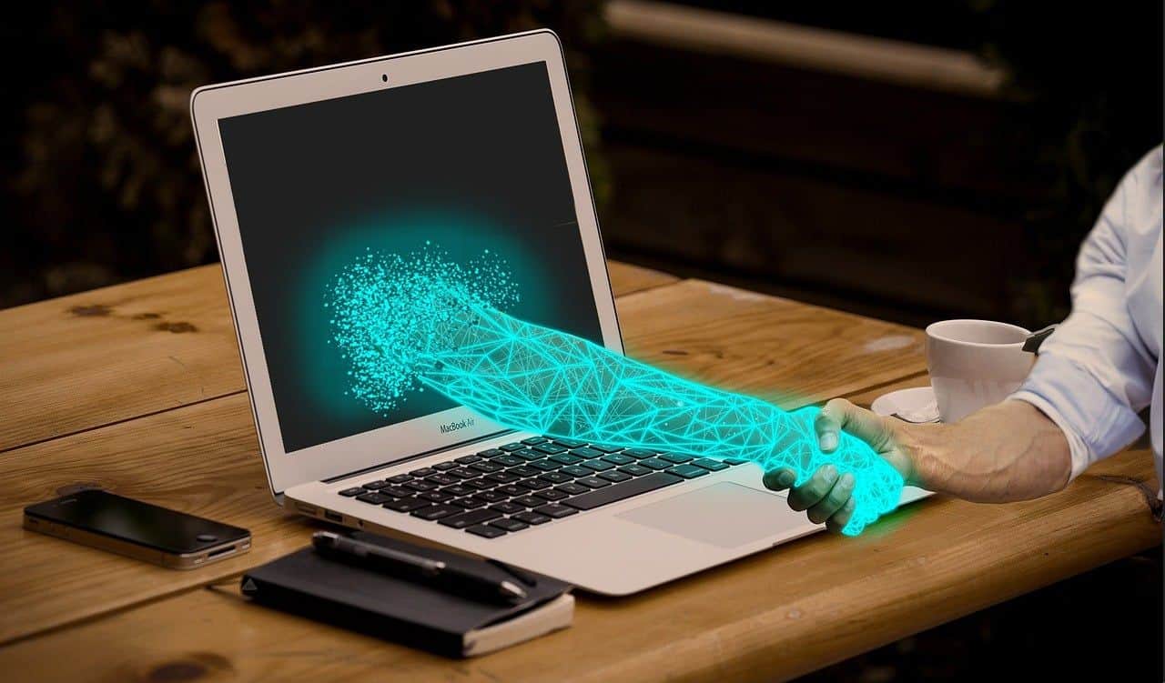Futuristic digital handshake between human and hologram on laptop screen.