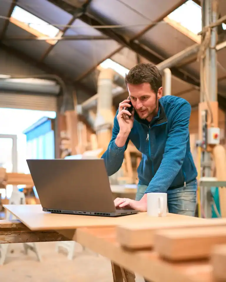 Man working on laptop in workshop, talking on phone, woodworking tools in background.