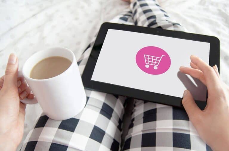 Person shopping online on tablet with shopping cart icon, coffee mug in hand.